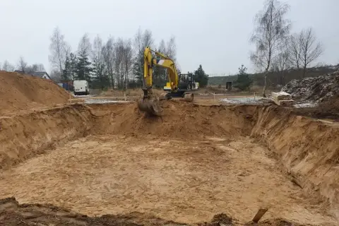 Żółta koparka pracuje przy wykopy pod fundamenty. W tle drzewa i biały van.